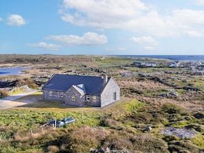A stone house with a sloped roof, surrounded by a rural landscape with a body of water and a small settlement in the distance.
