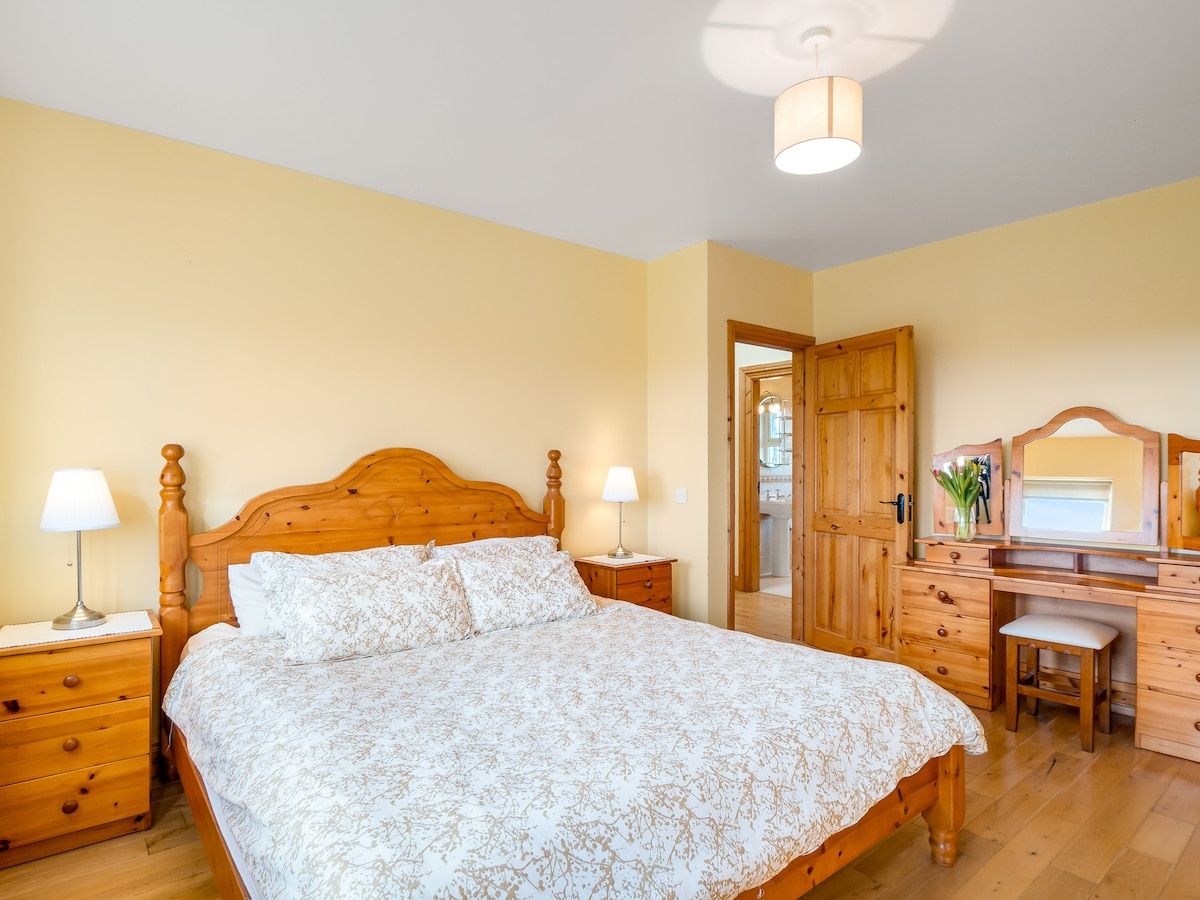 Ein Schlafzimmer mit einem hölzernen Bett, Nachttischen, einem Schreibtisch mit Spiegel und einem Sessel.