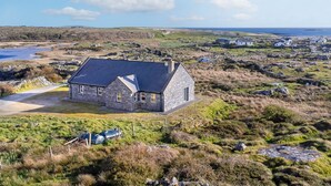 A stone house with a sloped roof, surrounded by a rural landscape with a body of water and hills in the background.