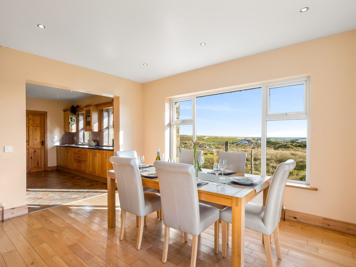 Un coin repas avec une table dressée pour quatre personnes, une cuisine en arrière-plan et une vue sur la campagne par de grandes fenêtres.