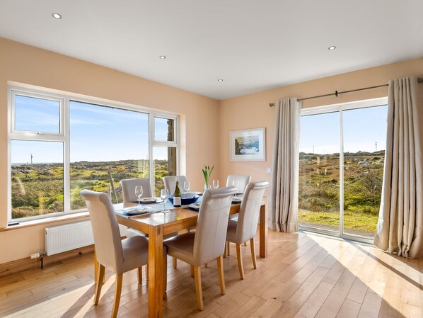 A dining room with a wooden table and chairs, large windows with a view of the countryside, and a radiator under the windows.
