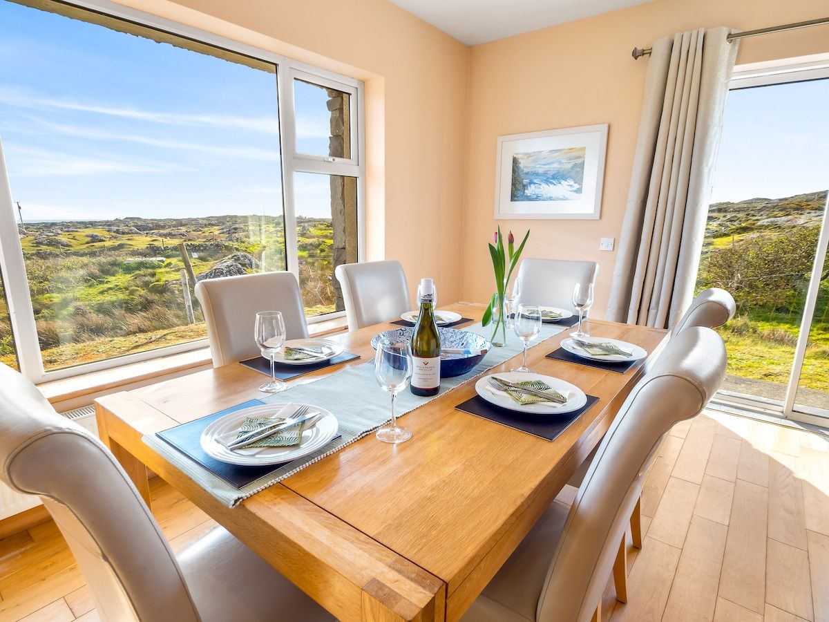 A dining table set for six with a bottle of wine, glasses, and plates, overlooking a scenic landscape.