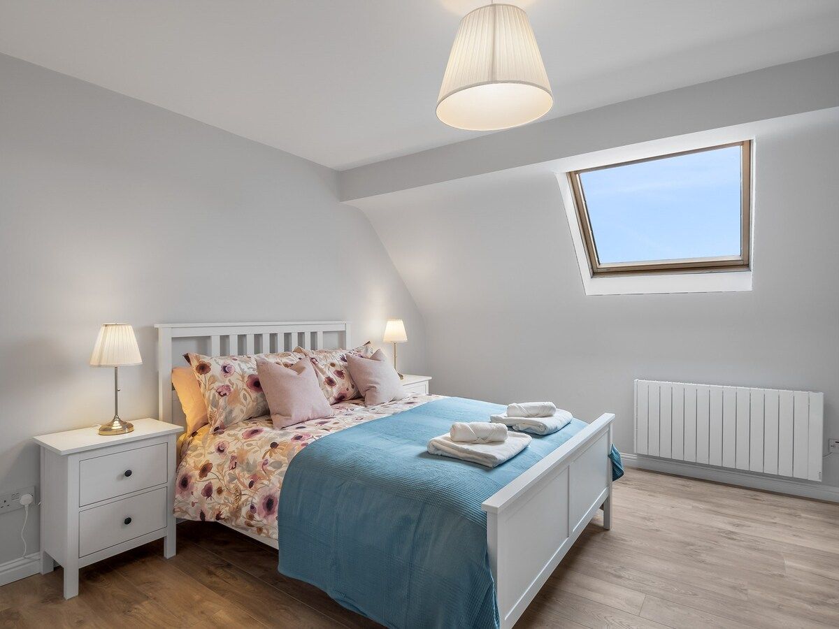Une chambre avec un plafond en pente, un puits de lumière, un lit recouvert d’une couvre-lit à motifs floraux, deux tables de chevet avec des lampes et un radiateur.
