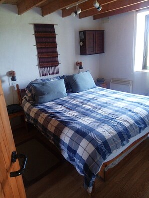 A bedroom with a bed, a nightstand, a lamp, a wall-mounted heater, and a window.