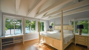 Uma cama com dossel e cabeceira de madeira, um banco perto da janela e uma mesinha de cabeceira com luminária.