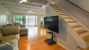 Uma sala de estar ampla com uma televisão de tela grande, piso de madeira, uma escada e vista através de janelas grandes.