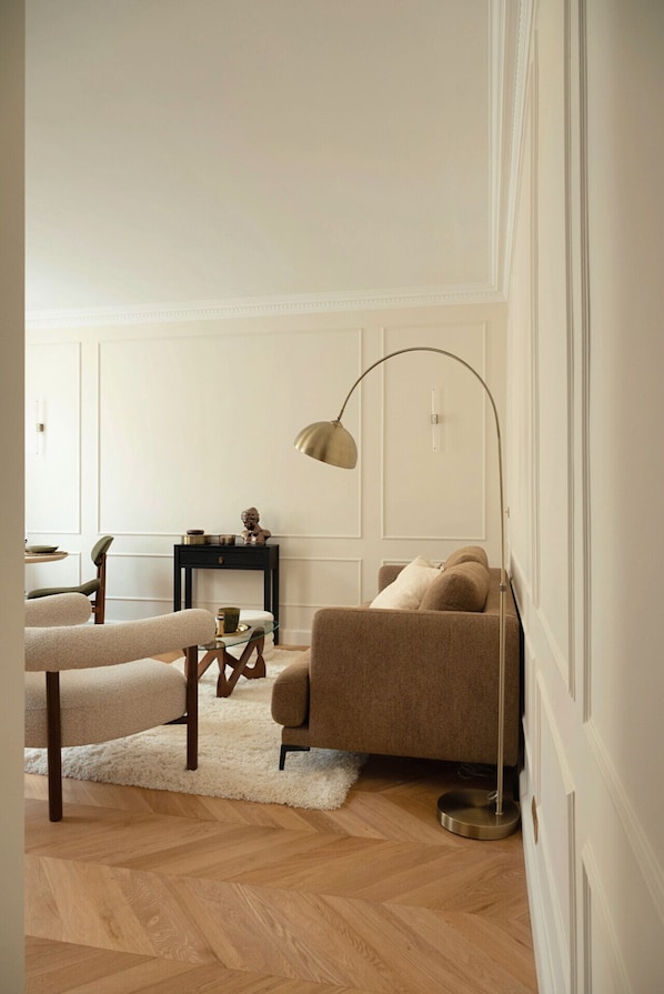 A living room with a sofa, a bench, a small table with decorative items, a floor lamp, and a herringbone-patterned wooden floor.