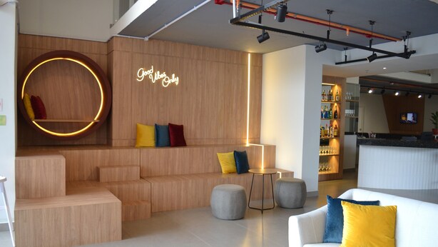 A modern lounge area with a wooden wall, a neon sign, and a bar in the background. - Ramada Encore Ponta Grossa  (Ponta Grossa)