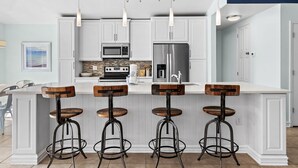 Une cuisine avec des meubles blancs, un îlot central doté de tabourets, un four à micro-ondes, un réfrigérateur et des luminaires suspendus.