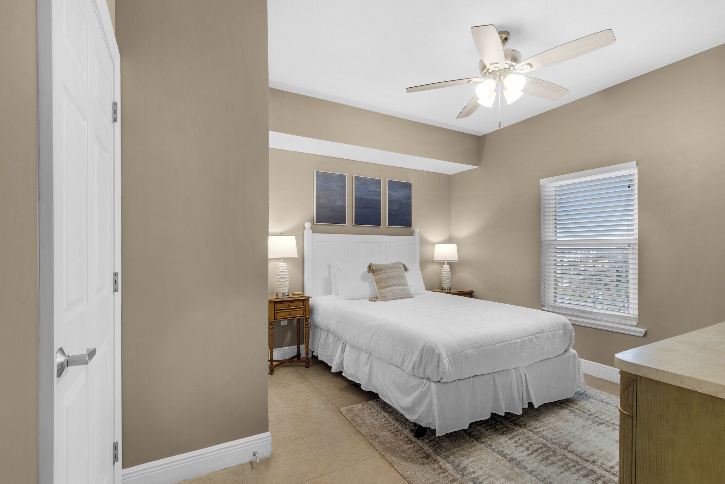 A bedroom with a bed, bedside lamps, a ceiling fan, a window with blinds, and a door.
