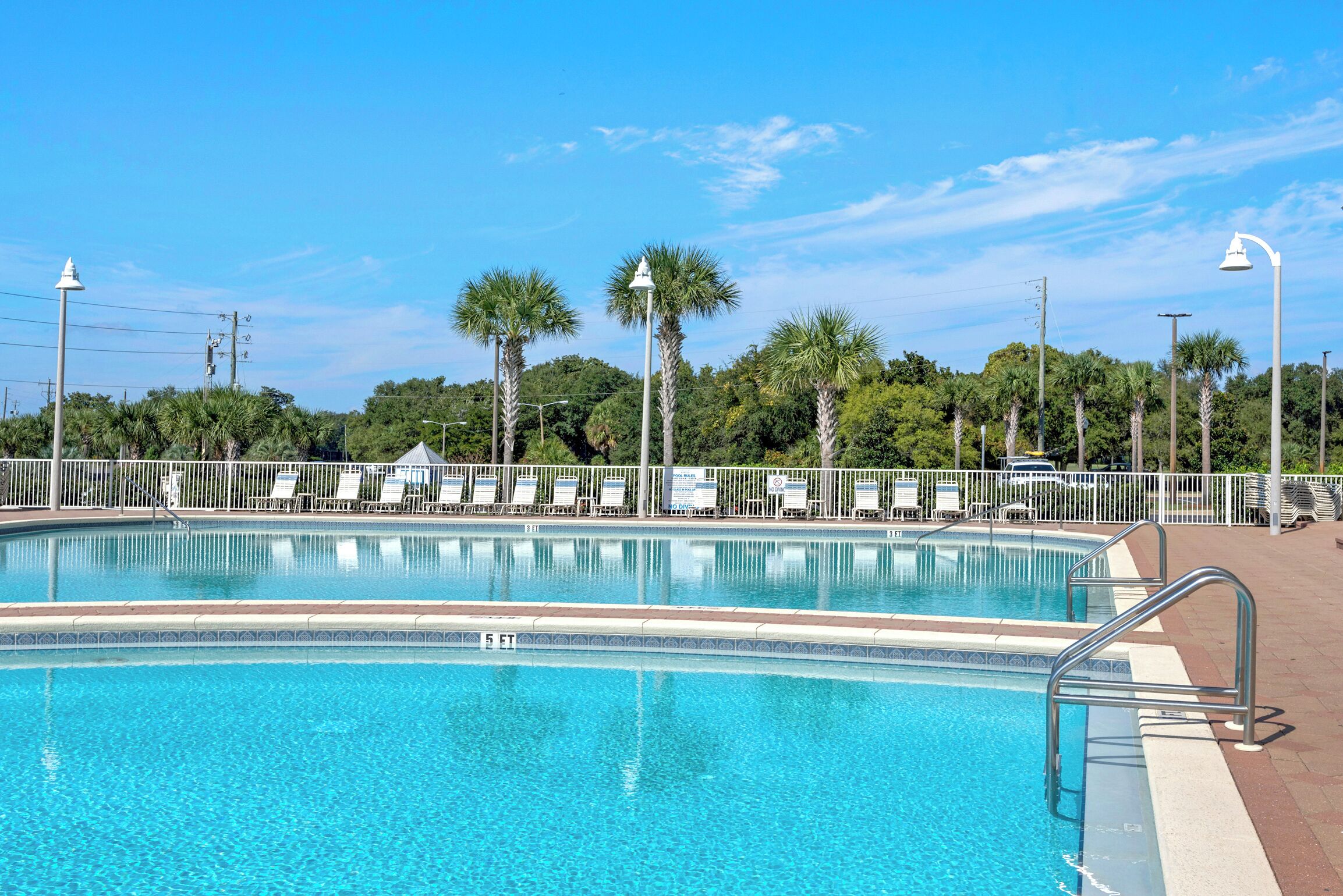 A clear blue swimming pool with a white fence, palm trees, and lounge chairs.