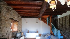Un saló rústic amb bigues de fusta al descobert, una paret de pedra, un sofà, una televisió i una escala amb barana blava.