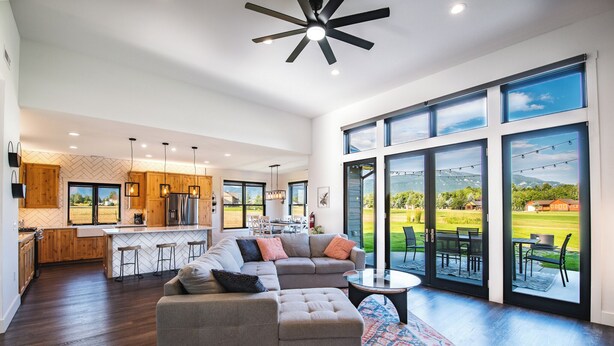 A spacious living room with a large sectional sofa, a coffee table, and a patterned rug. There is a kitchen area with wooden cabinets and a dining area with a bar. Large sliding glass doors lead to a patio with outdoor seating. - Luxurious Home in Red Lodge MT, Golf Course Retreat with Stunning Mountain Views (Red Lodge)