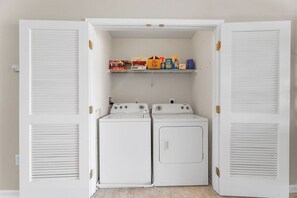 A laundry room with two white front-loading washers and dryers, a shelf with cleaning supplies, and a white door with louvered panels.
