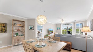 A dining area with a wooden table, chairs, and a chandelier. There is a sofa and a lamp in the background.