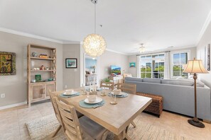 A dining area with a wooden table set for four, a chandelier, and a sofa in the background. - Sunset Island 6 Fountain Drive East, Unit 3B (Ocean City)