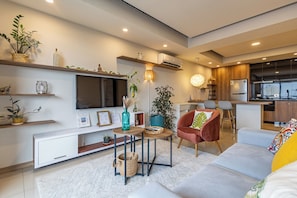 A modern living room with a flat-screen TV, a sofa, a red armchair, and a kitchen area.