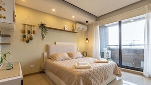 A modern bedroom with a large bed, a bedside table, a hanging plant, and a view of the city.