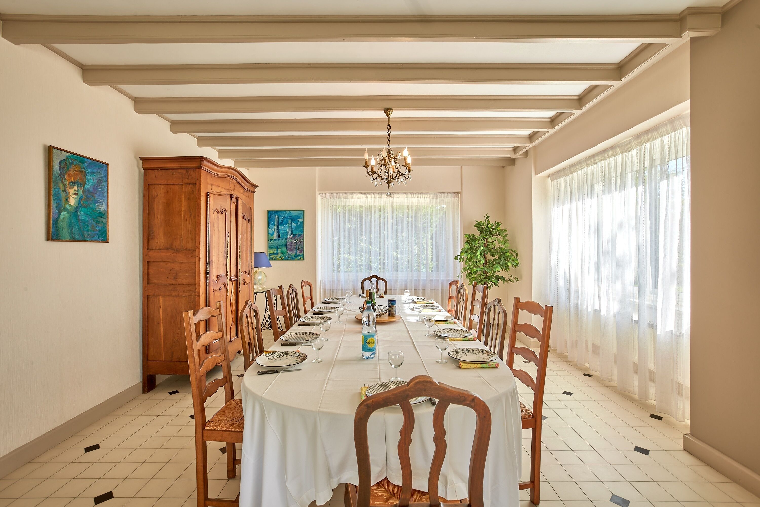 Een eetzaal met een lange tafel gedekt voor een maaltijd, houten stoelen, een kroonluchter en grote ramen met transparante gordijnen.