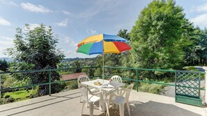 Eine Terrasse im Freien mit Tisch und Stühlen, einem bunten Sonnenschirm und Blick auf Grünflächen und Hügel.