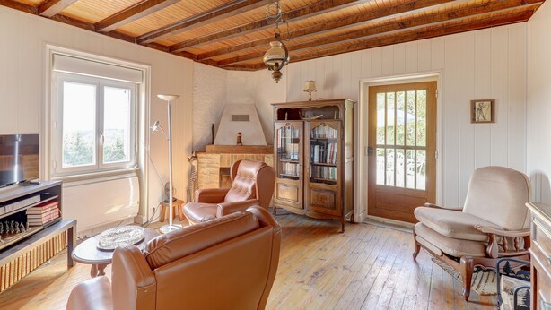 Ein geräumiges Wohnzimmer mit hoher Holzbalkendecke, Kamin, Ledersesseln, Bücherregal und Fernseher.