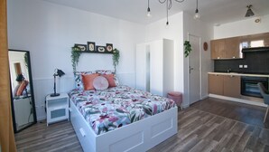 A modern bedroom with a large bed, bedside tables, a mirror, and a small desk. There is a kitchen area with cabinets and a countertop. - L'Etudiant - charmant studio (Saint-André-les-Vergers)