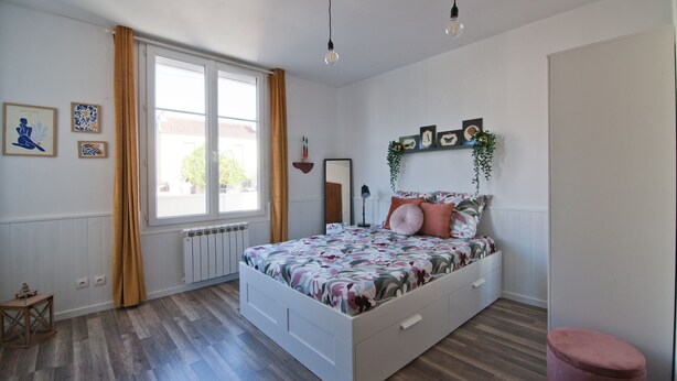 A bedroom with a large bed, a window with curtains, a radiator, and a small table. - L'Etudiant - charmant studio (Saint-André-les-Vergers)