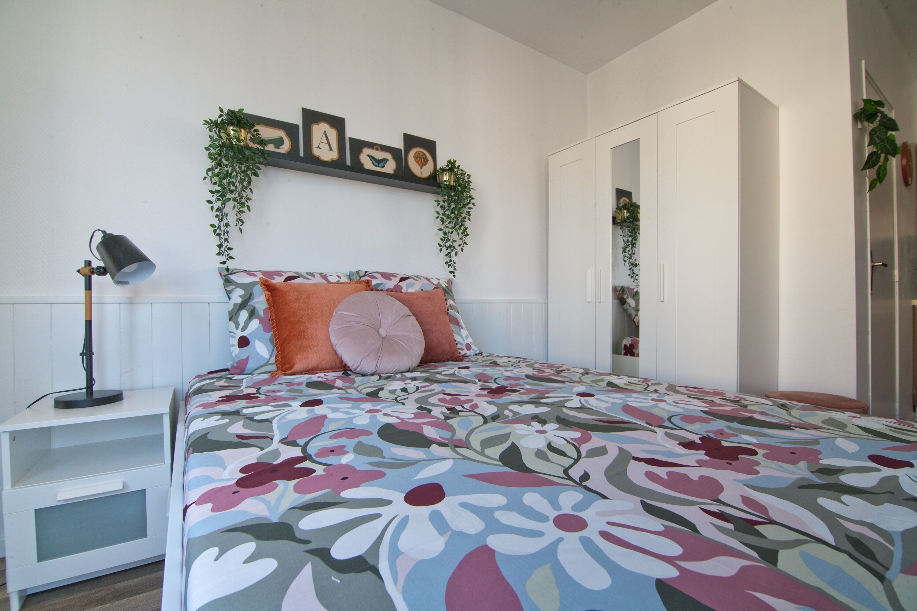 A bedroom with a floral bedspread, a bedside lamp, a wardrobe, and wall-mounted greenery.