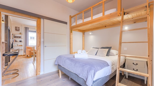 A bunk bed with a wooden ladder, a bedside table, and a chair in a well-lit room with wooden flooring. - Ernest - appt calme, proche centre de nantes (Nantes)
