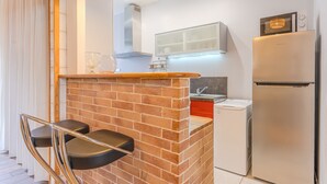 A compact kitchen with a brick accent wall, a stainless steel refrigerator, a washing machine, and a small countertop with a glass and a metal tray. - Ernest - appt calme, proche centre de nantes (Nantes)