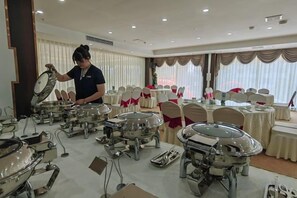 A banquet hall with round tables set for dining, a buffet area with food service equipment, and a person attending to the buffet setup. - Liancheng zhilong Hotel (Liancheng)