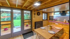 A wooden interior with a dining table set for four, a kitchen area with a built-in oven, and large sliding glass doors leading to a garden.