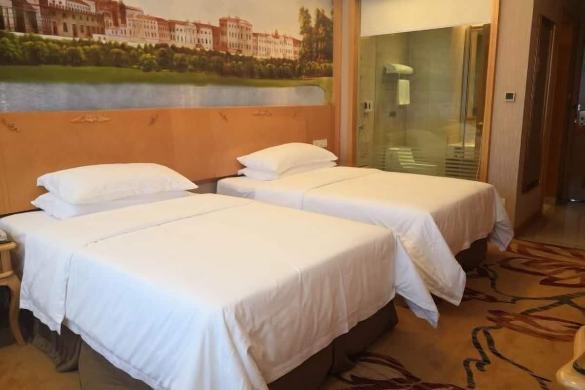 Quarto de hotel com duas camas de solteiro, cabeceira de madeira, um mural na parede e box de vidro para o chuveiro.