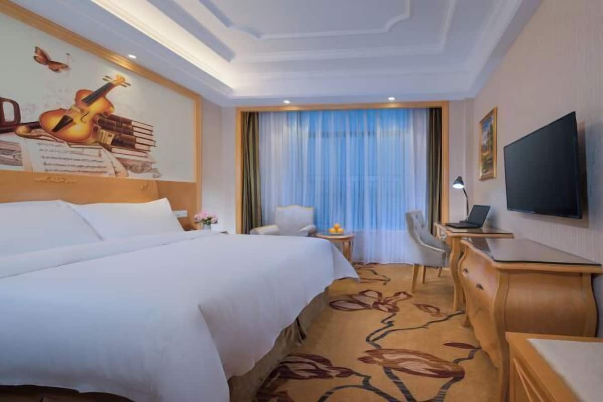 Quarto de hotel com uma cama grande, uma mesa com um laptop, uma televisão e uma janela com cortinas.