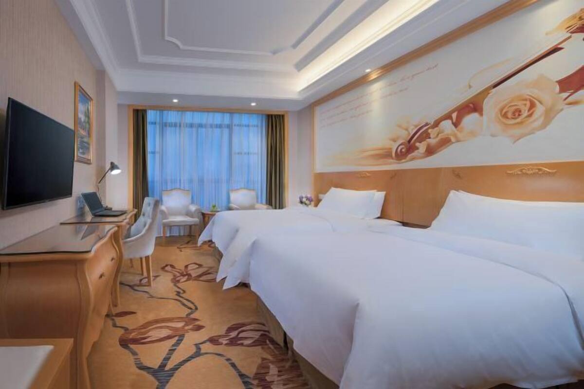 Quarto de hotel com duas camas, uma mesa de trabalho com televisão, uma cadeira e uma grande obra de arte floral na parede.