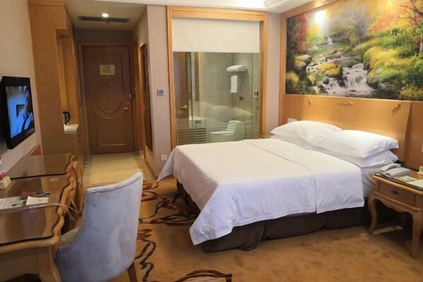 A hotel room with a bed, a desk, a chair, a TV, and a painting of a waterfall. - Vienna International Hotel (Zijin)