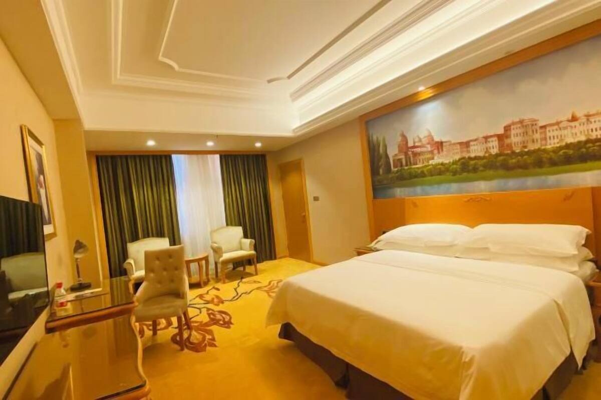 Quarto de hotel com uma cama grande, uma mesa de trabalho com cadeira, televisão e um quadro na parede.