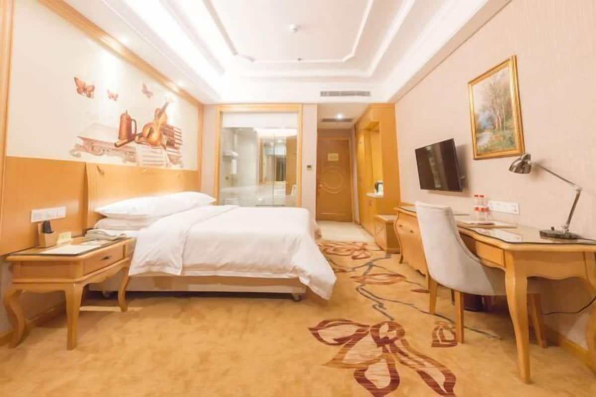 Quarto de hotel com cama, escrivaninha, cadeira, televisão e um quadro na parede.