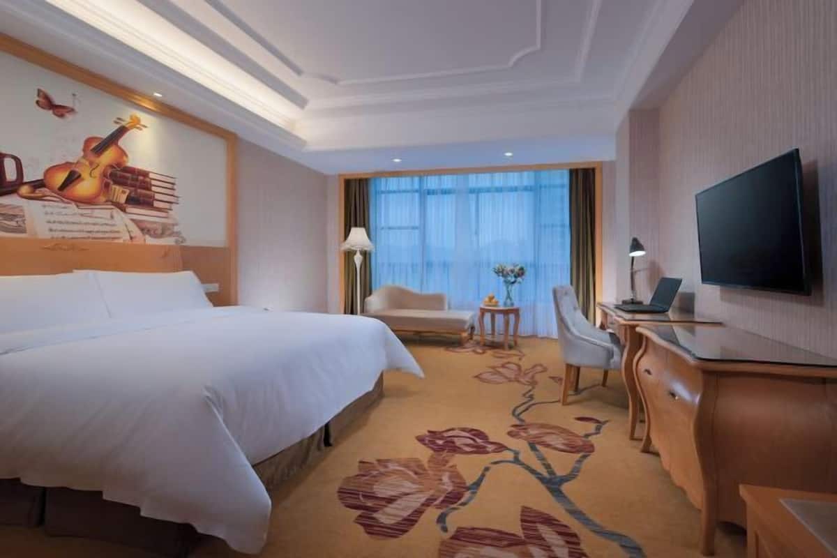 Quarto de hotel com uma cama grande, uma escrivaninha com computador, televisão, cadeira e janela com cortinas.