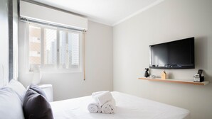 Une chambre d’hôtel avec un lit, un téléviseur fixé au mur, une fenêtre avec des stores et une table de chevet avec une lampe et une horloge.