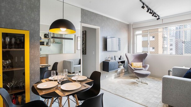 Un espace de salle à manger moderne avec une table dressée pour quatre personnes, un luminaire suspendu noir et une vue sur la cuisine et le salon, grâce à une porte ouverte.