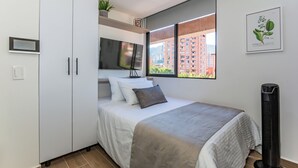 Un dormitorio con cama, televisor, ventana con vista a la ciudad y planta en la repisa.