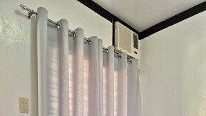 Un lit avec du linge de lit blanc et deux oreillers gris, un rideau blanc et un ventilateur de plafond.