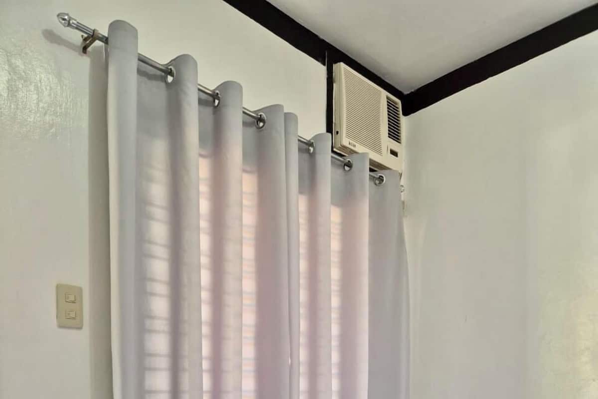En seng med hvide sengetæpper og to grå puder, et hvidt gardin og en ventilator i loftet.