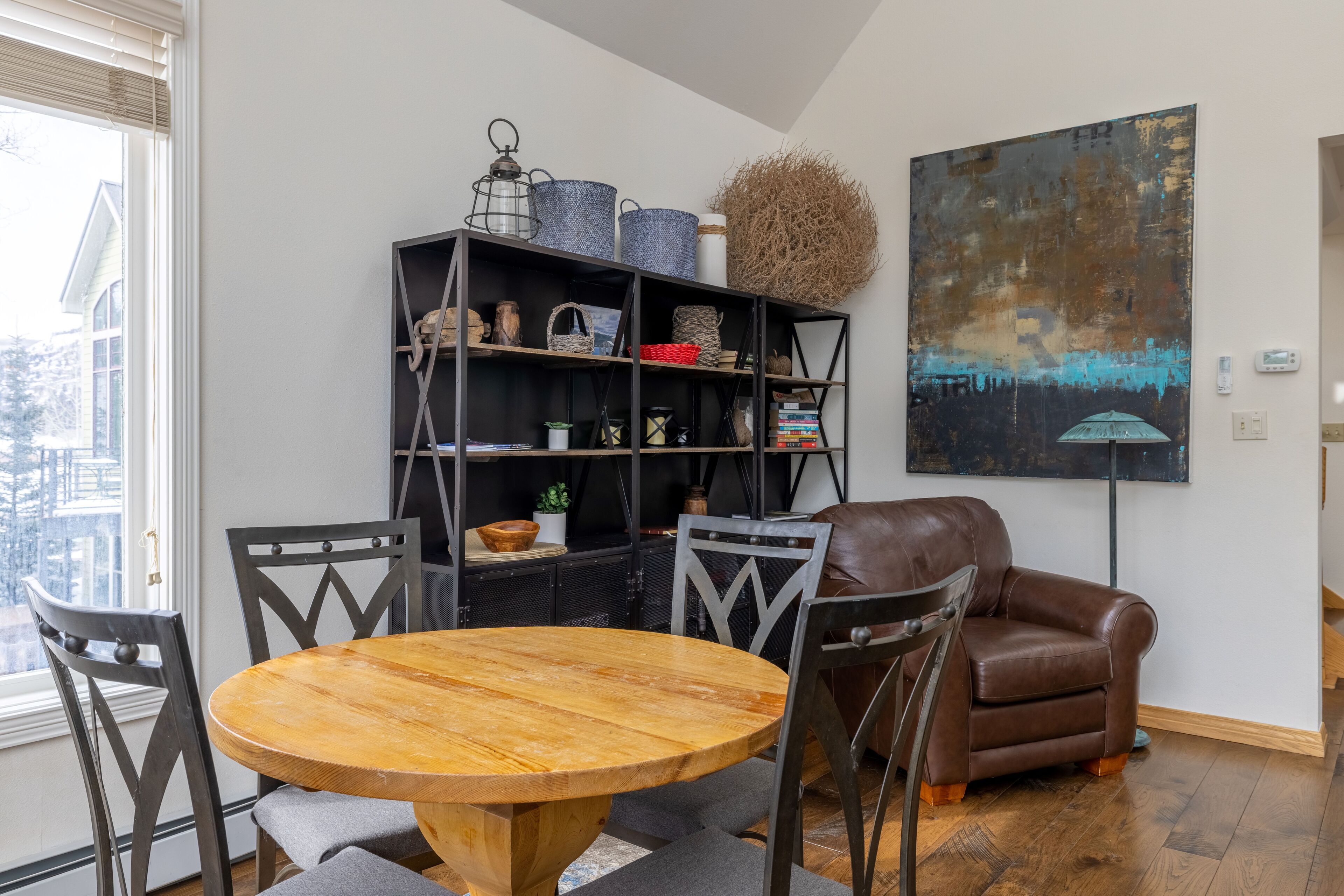 Ein Essbereich mit einem runden Holztisch und vier Stühlen, einem braunen Ledersofa, einem schwarzen Bücherregal mit verschiedenen Gegenständen und einem großen abstrakten Gemälde an der Wand.