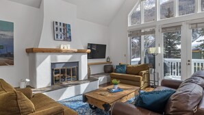 Un salon avec une cheminée, un téléviseur et une grande fenêtre donnant sur des arbres et une maison.
