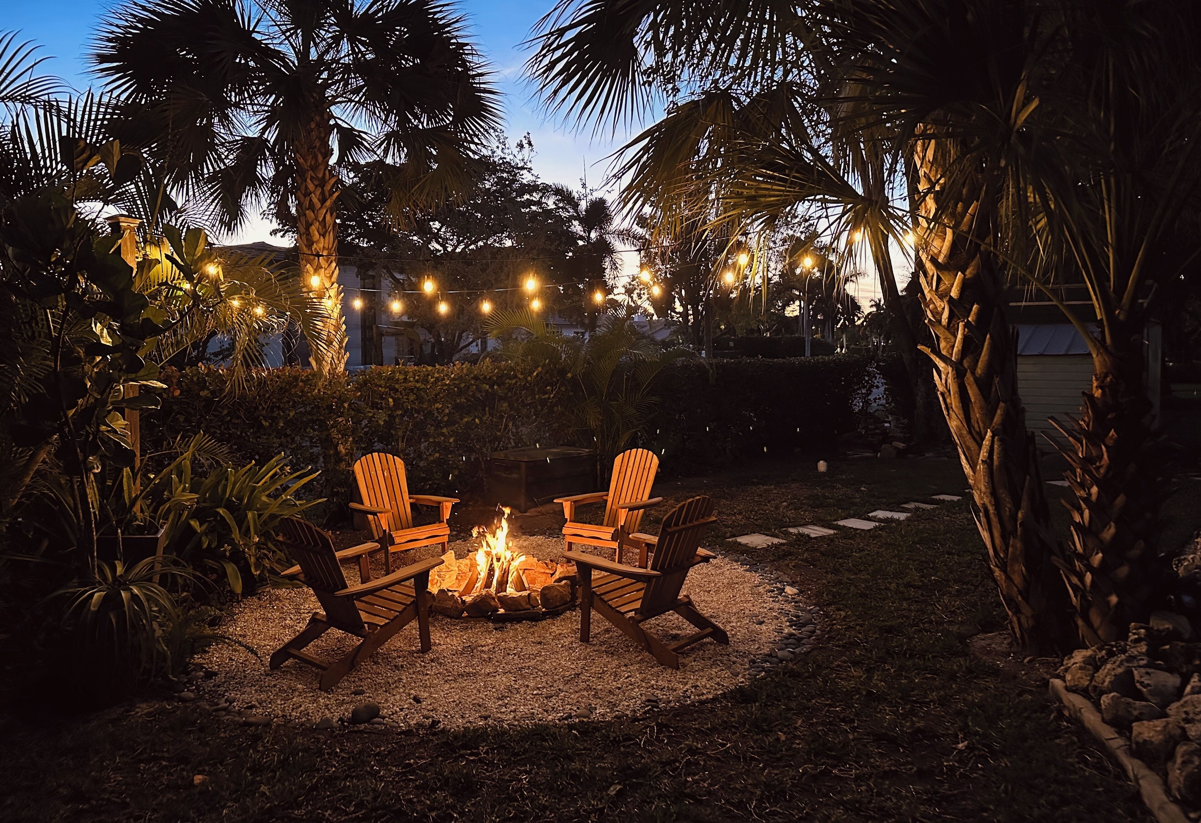 A fire pit with four Adirondack chairs arranged around it, surrounded by a gravel area with palm trees and string lights in the background.