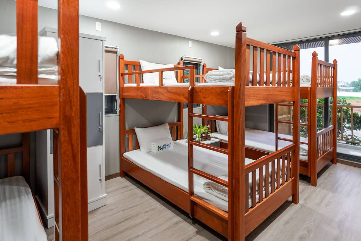 A bunk bed room with a wooden frame, a small bedside table with a plant, and a window with a view of greenery.