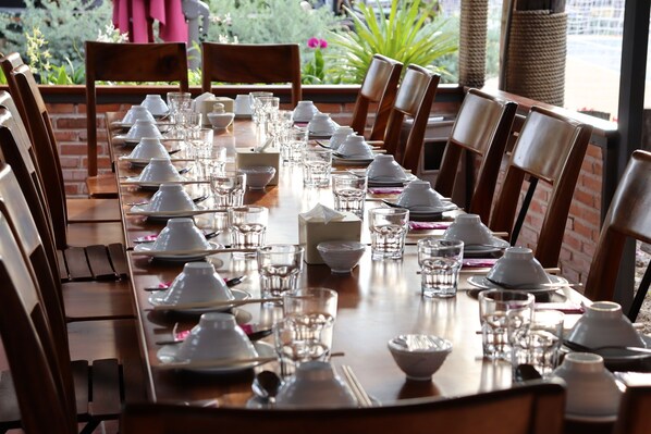 Una mesa de comedor lista para una comida, con platos, tazones y vasos blancos, rodeada de sillas de madera.