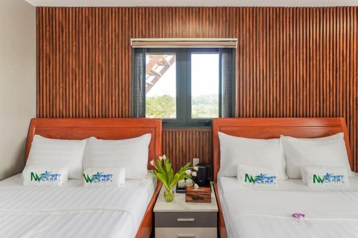 A double room with two beds, wooden paneling on the walls, a window with a view, and a nightstand with a vase of flowers.